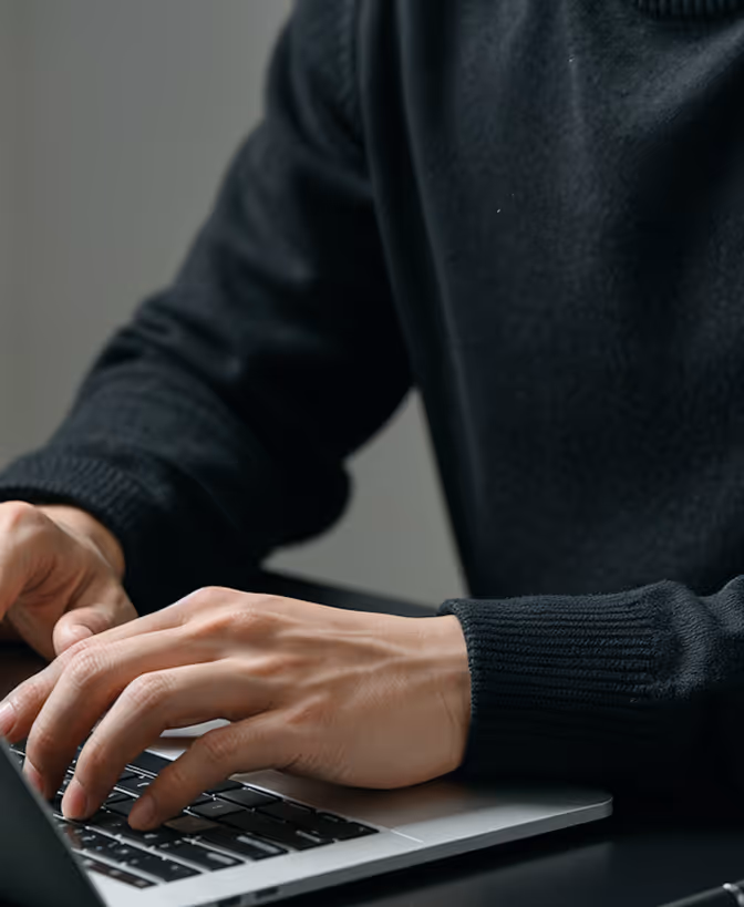 Pessoa de suéter preto digitando no teclado de um notebook em uma mesa escura.