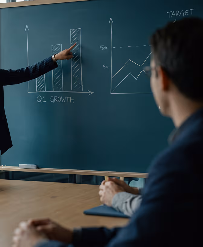 Pessoa apontando para um gráfico de barras em um quadro-negro mostrando o crescimento do Q1 enquanto outra pessoa observa.