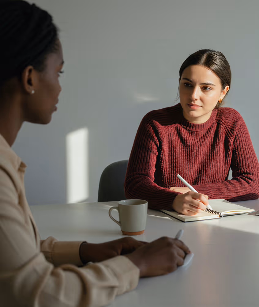 Duas mulheres sentadas em uma mesa em uma conversa séria, uma anotando em um caderno.