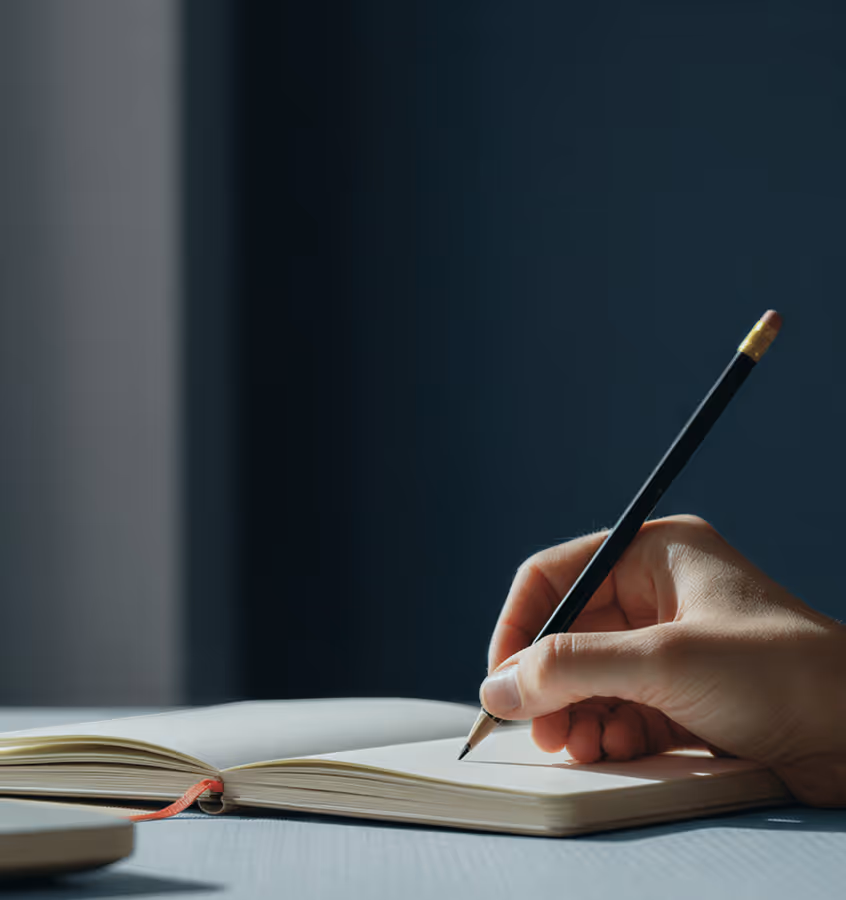 Mão segurando um lápis pronto para escrever em um caderno aberto sobre uma mesa com iluminação suave.
