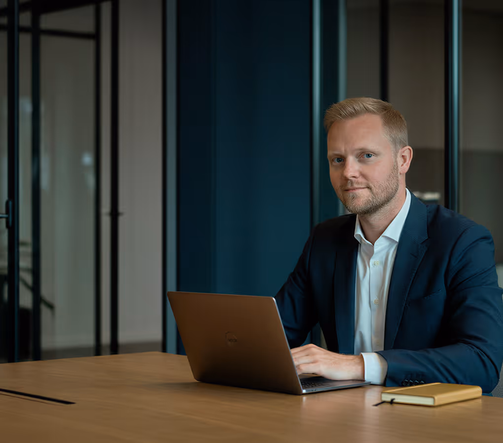 Homem de terno sentado em uma mesa usando um notebook com um caderno por perto em um escritório moderno.