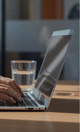 Pessoa digitando no teclado de um notebook com um copo de água sobre uma mesa de madeira.