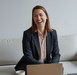 Mulher sorridente de blazer preto sentada em um sofá, com um notebook e uma xícara de café por perto.