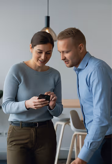 Dois colegas sorrindo e olhando juntos para um smartphone em um ambiente de escritório moderno.
