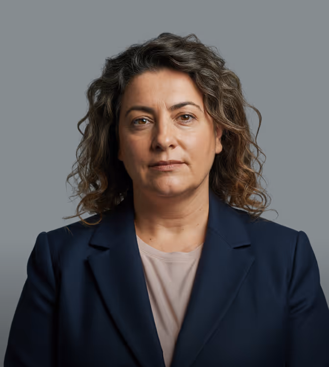 Mulher séria de meia-idade com cabelo cacheado usando blazer azul-marinho e camisa clara sobre fundo escuro.