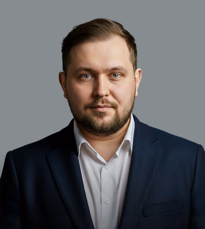 Retrato de um homem de cabelo curto e barba usando camisa branca e blazer escuro contra fundo preto.