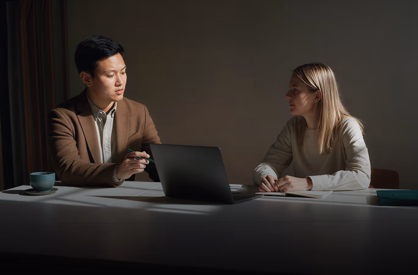 Duas pessoas sentadas em uma mesa conversando com um notebook e um caderno sob uma iluminação suave.