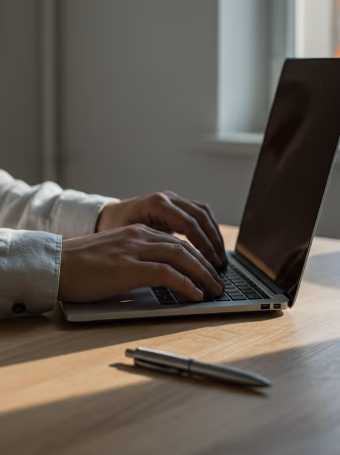 Pessoa digitando no teclado de um notebook em uma mesa de madeira com uma caneta por perto.