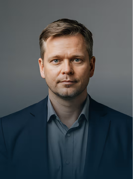 Retrato de um homem com cabelo castanho claro curto e olhos azuis, vestindo um blazer escuro e camisa cinza.