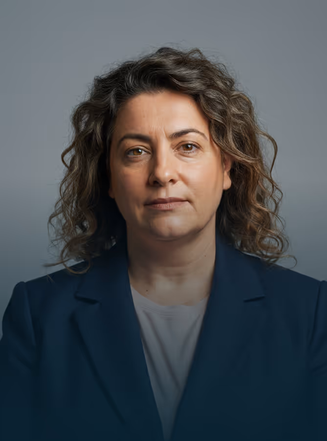 Mulher com cabelo cacheado, vestindo um blazer azul-marinho e camisa clara em fundo cinza.