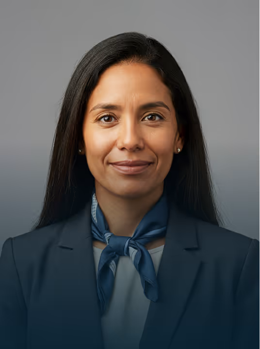 Mulher sorridente com cabelo preto longo, vestindo um blazer escuro e lenço azul no pescoço, em fundo cinza.
