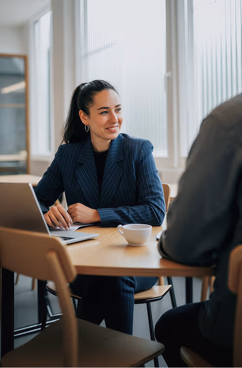 Mulher de blazer listrado sorrindo e conversando durante uma reunião em uma mesa com notebook e xícara de café.