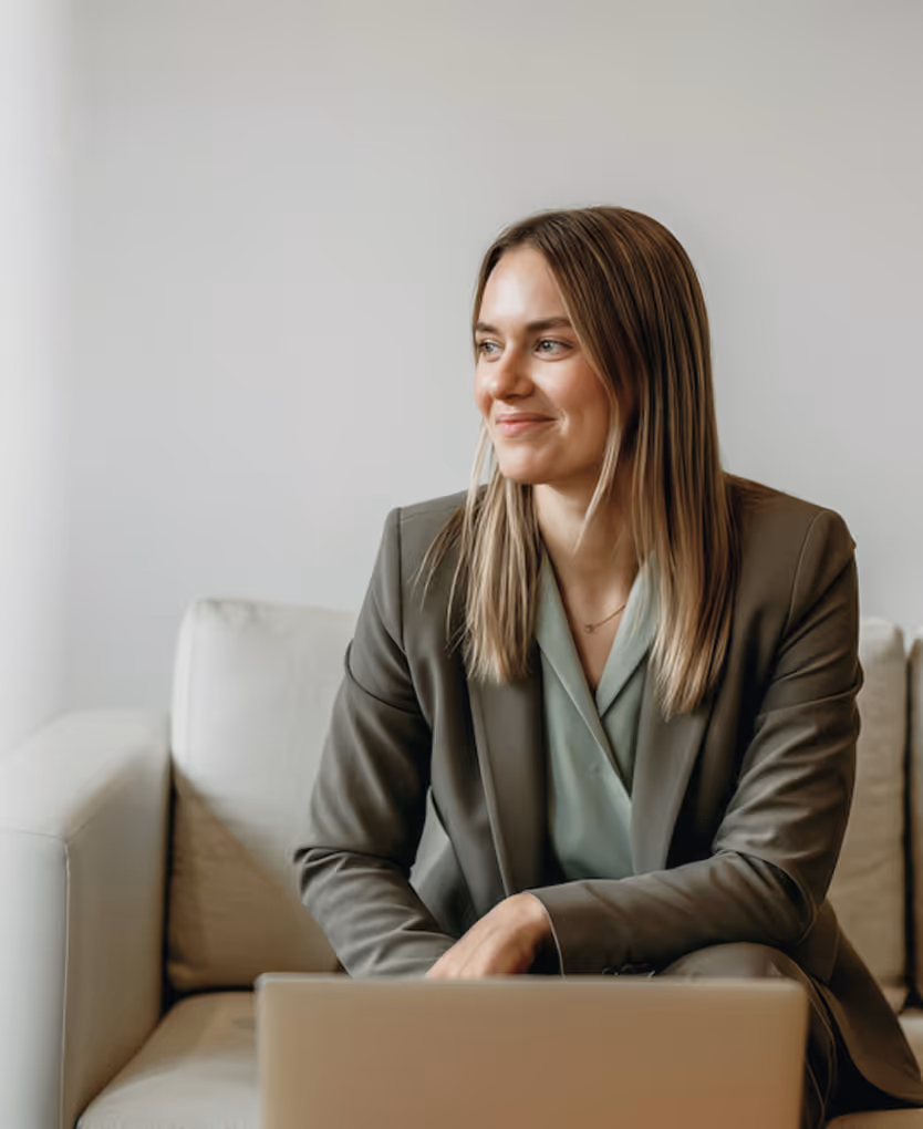 Mulher sorridente em traje de negócios sentada em um sofá com um notebook à frente, olhando para o lado.
