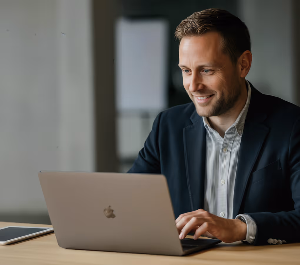 Homem sorridente de blazer digitando em um notebook em uma mesa de madeira com um tablet por perto.
