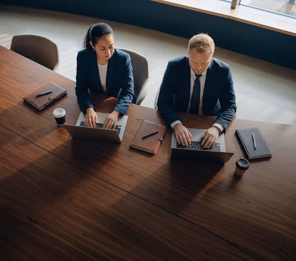 Dois profissionais digitando em notebooks lado a lado em uma mesa de confer&ecirc;ncia.