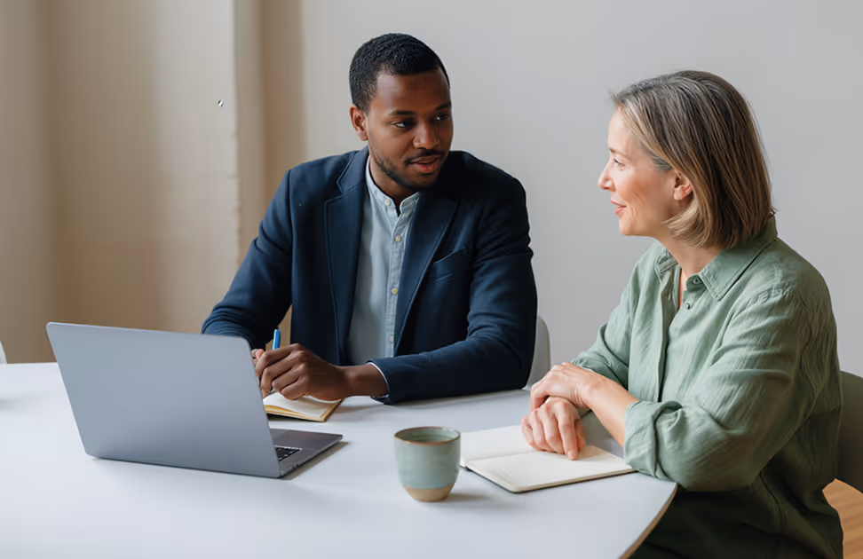 Duas pessoas sentadas em uma mesa conversando, com um notebook, um caderno e uma caneca de café à frente.
