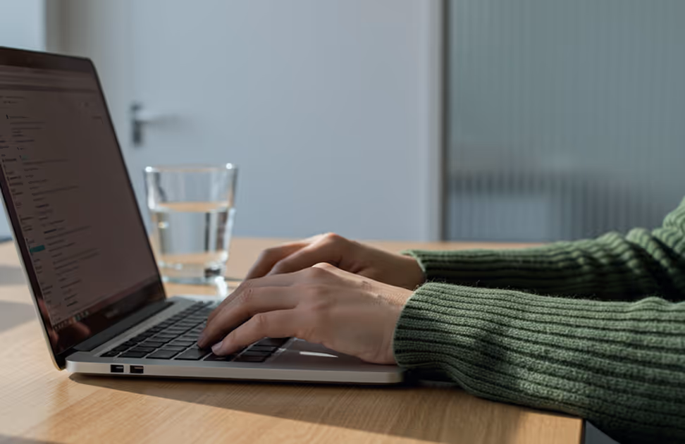 Pessoa digitando no teclado de um notebook com um copo de água sobre uma mesa de madeira por perto.