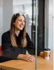 Mulher sorridente sentada em uma mesa com um copo de café para levar em um ambiente interno claro.