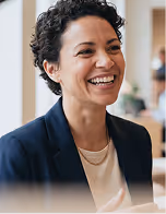 Mulher sorridente de cabelo curto e cacheado usando um blazer azul-marinho e camisa clara em ambientes internos.